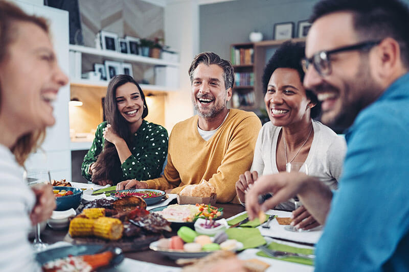 A group of five adults sits around a dining table, laughter ringing out as they enjoy a meal together. With hearing aids aiding their conversations, the table is spread with delicious dishes like corn on the cob and meats, nurturing a warm and friendly atmosphere.